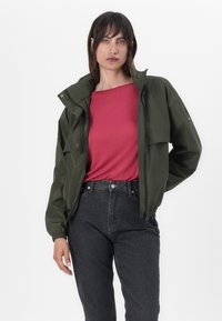 Casaco à prova de água verde-azeitona com fecho de correr, dois grandes bolsos no peito e punhos elásticos, usado sobre uma camisa rosa e jeans escuros.