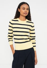 Žltý pruhovaný sveter s námorníckymi horizontálnymi líniami, rebrovaná textúra a okrúhly výstrih. Kombinovaný s tmavými nohavicami pre kontrastný vzhľad.