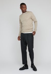 Pulover tricotat bej cu guler rotund, cu un finisaj texturat, asortat cu pantaloni negri și pantofi slip-on negri. Modelul stă pe un fundal neutru.
