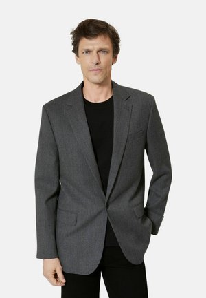 Homme portant un blazer gris à un bouton sur une chemise noire, debout avec une main dans la poche sur un fond clair uni.