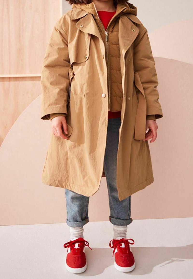 Enfant portant un trench-coat beige par-dessus une doudoune camel, un jean retroussé, des chaussettes blanches et des baskets rouges, debout sur un sol clair.