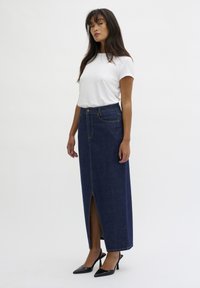 Gonna lunga in denim scuro con uno spacco anteriore, abbinata a una T-shirt bianca semplice. Presenta cuciture dorate e un design aderente. Tacchi alti neri.