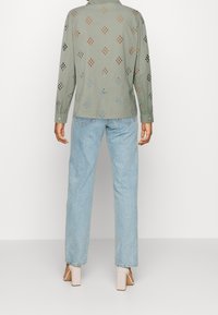 Groene shirt met lange mouwen en een patroon van uitgesneden diamanten in zwart, oranje en blauw. Gecombineerd met lichtblauwe recht-zittende jeans en beige hakken.