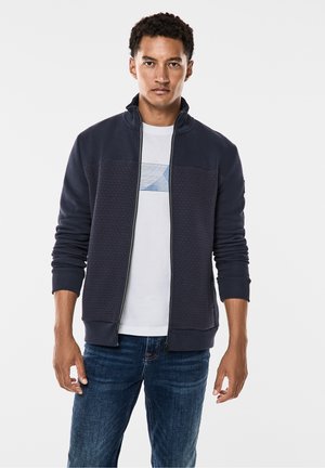 Marineblå lynlåsjakke med tekstureret panel, høj krave og ribbede manchetter, båret over en hvid T-shirt med blå grafik, parret med jeans.