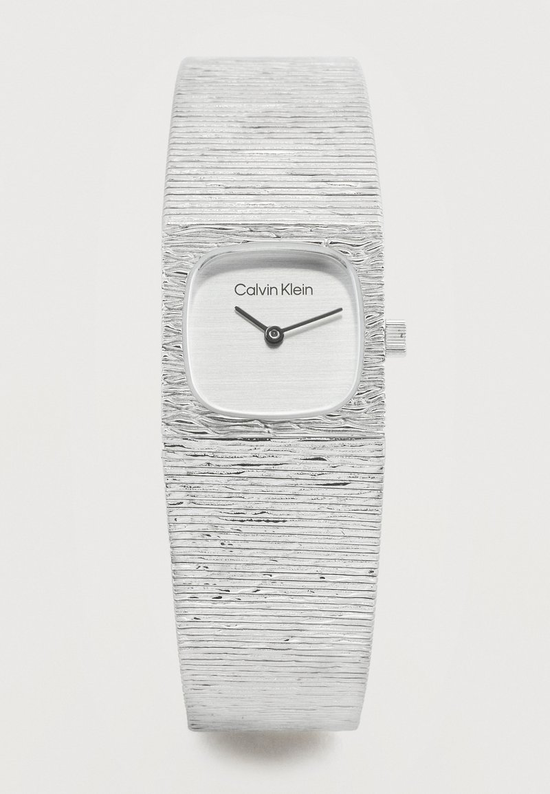 Ασημένιο ρολόι χειρός Calvin Klein με υφασμένο φαρδύ λουράκι και μικρό τετράγωνο καντράν που δείχνει ώρα 10:10.