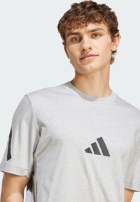 Šedé bavlněné tričko s kulatým výstřihem a krátkými rukávy, na hrudi nese černé grafické logo a další na levém rukávu.