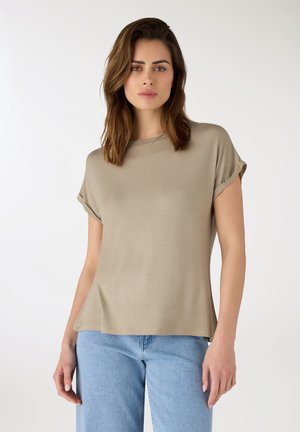 Donna con una maglietta beige a maniche corte con risvolti e jeans azzurri, in piedi davanti a uno sfondo bianco.