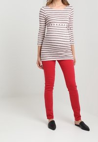Blusa de maternidad con cuello barco, rayas horizontales en gris y blanco, combinada con unos jeans skinny rojos. La modelo lleva puestas unas zapatillas negras sin cordones.