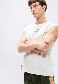 Λευκό αμάνικο μπλουζάκι με εφαρμοστή σχεδίαση, που διαθέτει μαύρο γραφικό στο στήθος και κόκκινη ετικέτα στο πλάι. Ομαλή υφή υφάσματος.