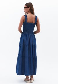 Denim-maksimekko, jossa on smockattu yläosa, röyhelöolkaimet ja kerroksellinen helma. Ominaisuuksiin kuuluu syvä sininen väri ja pehmeä rakenne.