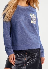Sudadera azul de tejido suave con cuello redondo, que presenta un parche de letra bordado y colorido en el lado izquierdo del pecho. Combinada con una falda negra de cuero sintético.