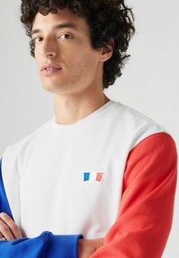 Witte sweatshirt met blauwe en rode mouwen, met een klein geborduurd ontwerp dat lijkt op de Franse vlag op de borst. Gladde textuur.
