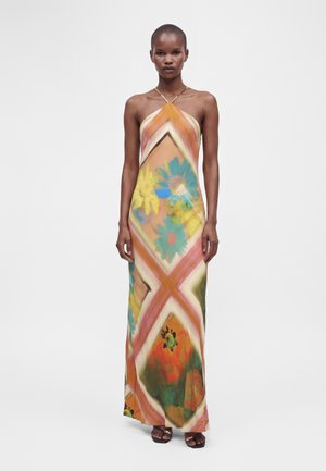 Femme portant une robe sans manches longue jusqu'au sol avec de grands motifs floraux et géométriques colorés, debout devant un fond blanc uni.