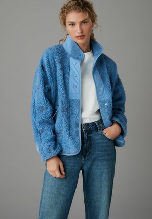 Giacca di fleece blu con tessuto testurizzato, bottoni a scatto e colletto blu a contrasto, abbinata a jeans di denim azzurro chiaro.