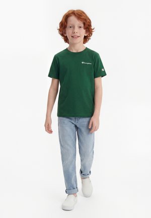 Rødhåret dreng iført en grøn Champion t-shirt, lyseblå jeans med manchetter og hvide slip-on sko, går fremad på en hvid baggrund.