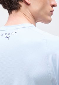 Νεαρό άτομο που φοράει ανοιχτό μπλε αθλητικό πουκάμισο με τρύπες, με τα λογότυπα "HYROX" και Puma στο πάνω μέρος της πλάτης, κοιτάζοντας προς τα δεξιά.