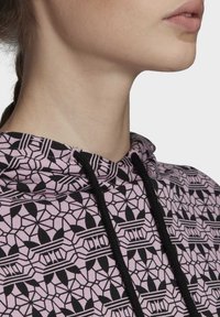 Mikina s růžovým vzorem a geometrickými černými motivy. Obsahuje šňůrku a texturovanou látku. Detailní záběr ukazuje výstřih.