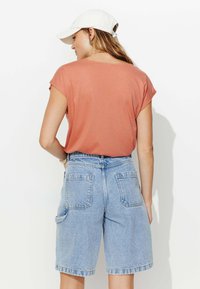 T-shirt couleur rouille à manches courtes associé à un short en jean bleu clair avec deux poches arrière. Matière en coton avec une coupe décontractée.