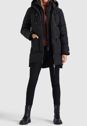 Mujer con abrigo acolchado negro con capucha y múltiples bolsillos con cremallera, pantalones negros, botas negras con cordones y suéter de cuello alto color óxido.