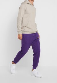 Beige mikina s kapucí a přední kapsou, zdobená zlatou výšivkou, ve spojení s fialovými tepláky s odpovídajícím logem. Bílá sneakers dotváří celý outfit.