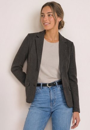 Femme aux cheveux attachés, portant un blazer sombre, un haut beige, un jean bleu et une ceinture noire, debout devant un fond clair uni.