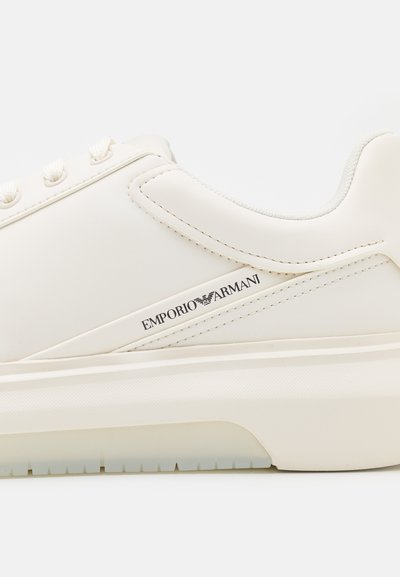 Bela usnjena športna obutev s gladko teksturo, vidnim blagovnim znakom in oblazinjeno podplatko. Ima minimalističen oblikovanje in teksturirane poudarke.