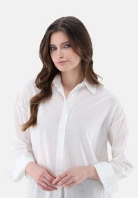 Blusa bianca e trasparente con motivo a pois, maniche lunghe, chiusura frontale a bottoni e colletto classico; il tessuto sembra leggero e testurizzato.