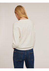 Sudadera de color crema con un corte holgado, hombros caídos y puños acanalados, combinada con unos vaqueros de mezclilla azul oscuro que tienen bolsillos traseros.