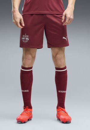 Mann trägt dunkelrote Red Bull Salzburg Fußballshorts und Socken mit leuchtend orangefarbenen Fußballschuhen und steht vor grauem Hintergrund.