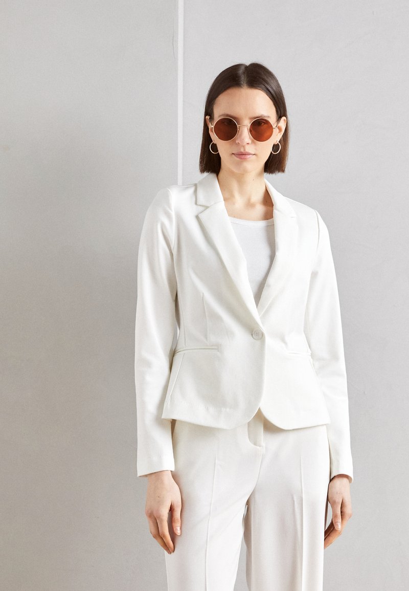 Weißer taillierter Blazer mit einer figurbetonten Passform, ein Knopf und zwei vorderen Taschen. Kombiniert mit passender Hose und runden Sonnenbrillen.