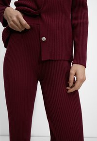 Burgundowy zestaw swetra i spodni z prążkowanego materiału, prezentujący teksturowaną tkaninę, długie rękawy i guziczek na górze.