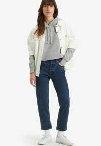 Giacca di jeans bianca sopra un hoodie grigio, jeans blu a vita alta e sneakers bianche con finitura testurizzata. Design minimalista, vestibilità casual.