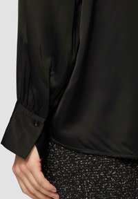 Black satin blouse with a loose fit, gathered sleeve cuffs, and a button detail at the wrist, paired with a textured black skirt.
