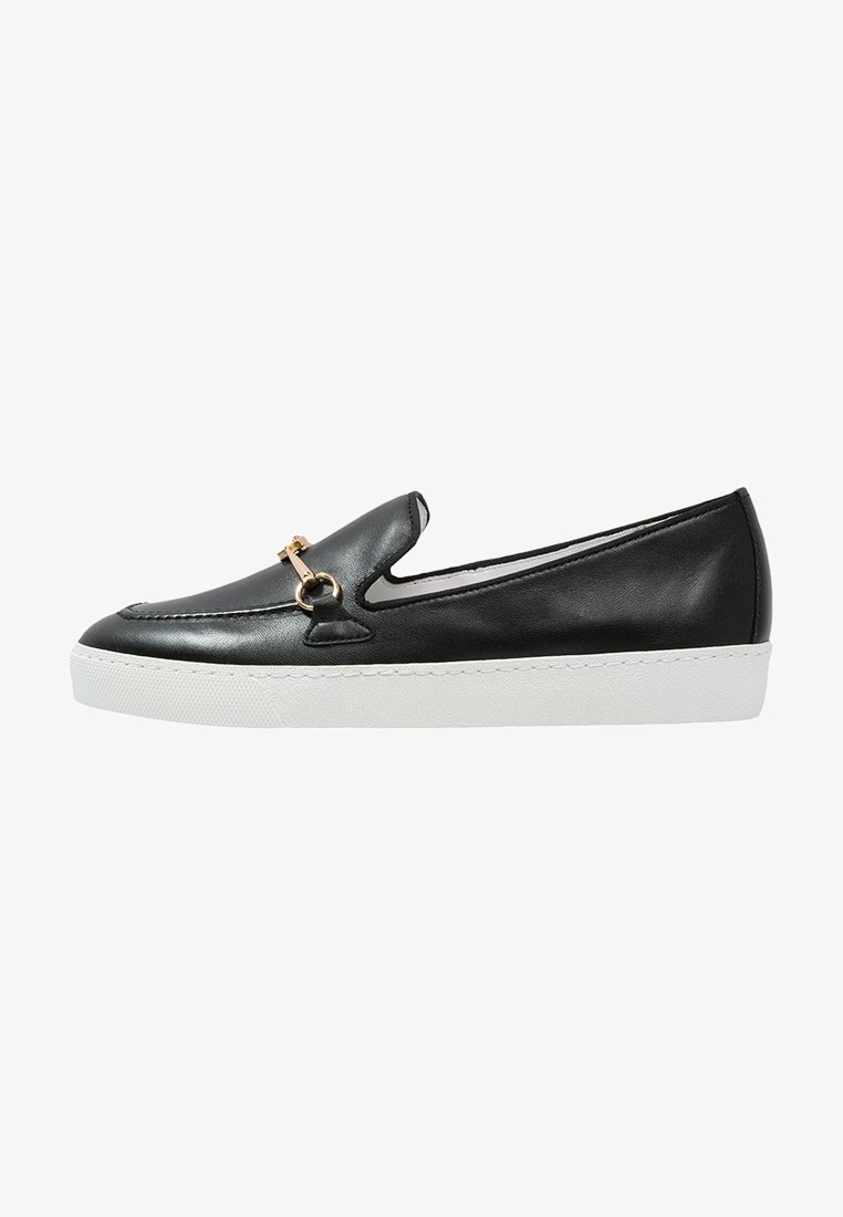 Chaussure noire en cuir à enfiler avec une semelle en caoutchouc blanche. Présente un ornement en métal doré décoratif sur le cou-de-pied et une texture lisse.