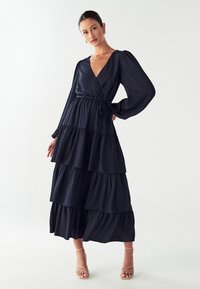 Dolga mornarsko modra obleka s plasteh in ovito zasnovo, z V-izrezom in napihnjenimi rokavi. Narejena je iz lahkotnega materiala z gladko teksturo.