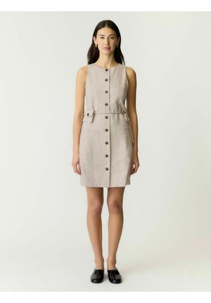 Femme debout portant une robe beige sans manches avec boutons, poches latérales et des chaussures plates noires sur un fond clair uni.
