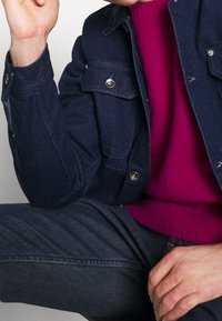 Giacca di jeans blu scuro con bottoni argentati, sovrapposta a un maglione a maglia fucsia brillante, indossata con jeans scuri. Mano posizionata in modo riflessivo.