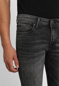 Jeans gris oscuro con un patrón de desvanecimiento, diseño de cinco bolsillos y cierre de botón metálico. La tela parece denim con una textura suave.