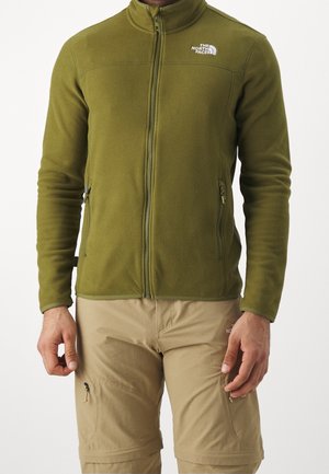 Mann trägt olivgrüne Fleecejacke mit Reißverschluss und zwei Reißverschlusstaschen und beige Cargohosen, steht vor einem einfarbigen Hintergrund.