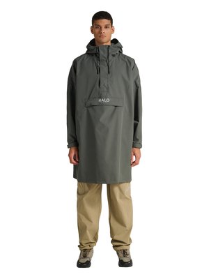 Jonge man staat, draagt een donkergroene lange regenjas met capuchon en de tekst "HALO" en beige broek met sneakers, kijkt recht vooruit tegen een witte achtergrond.