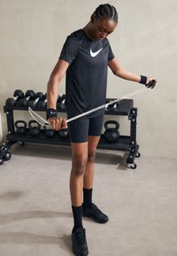 Črn Nike majica s belim logotipom, ujemajoče črne kratke hlače in črne atletske čevlje. Oseba uporablja odpornostne bande v telovadnici.