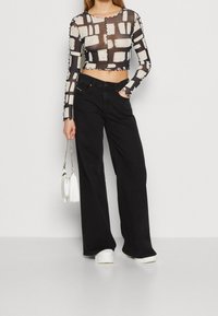 Nainen, jolla on päällä mustavalkoinen kuvioitu läpikuultava crop-top, mustat leveälahkeiset farkut, valkoiset lenkkarit ja pieni valkoinen käsilaukku.