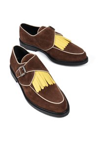 Brązowe zamszowe buty z żółtym frędzlem, czarną podeszwą i dekoracyjnym paskiem z klamrą, podkreślone kremowym wykończeniem.