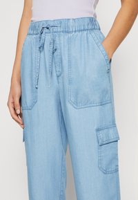 Jasnoniebieskie dżinsowe spodnie cargo z sznurkiem w pasie, kieszeniami po bokach oraz naszywanymi kieszeniami na obu nogawkach. Miękki, lekko teksturowany materiał.