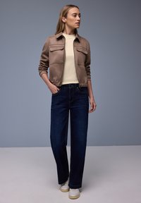 Femme debout les mains dans les poches, portant une veste zippée marron clair, une chemise crème, un jean bleu foncé et des baskets blanches sur fond gris.