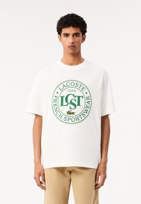 Camiseta blanca de algodón de manga corta con un logotipo circular verde de Lacoste, el texto "LST PARIS FRENCH SPORTSWEAR" y un pequeño gráfico de cocodrilo.