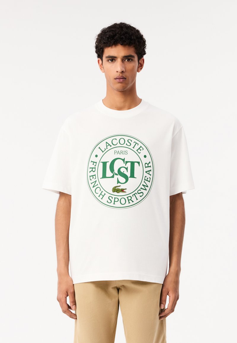Camiseta blanca de algodón de manga corta con un logotipo circular verde de Lacoste, el texto "LST PARIS FRENCH SPORTSWEAR" y un pequeño gráfico de cocodrilo.
