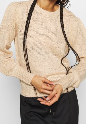 Personne portant un pull en maille beige à manches longues et un pantalon noir, les mains jointes à la taille, avec de longs cheveux tressés sur le pull.