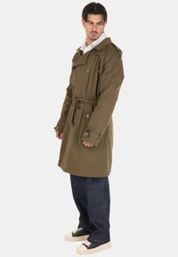 Cappotto trench verde oliva con bottoni doppi, polsini regolabili e vita con cintura, abbinato a pantaloni in denim scuro e scarpe nere.