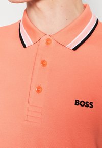 Polo à manches courtes en coton corail avec une finition texturée, trois boutons orange et un col rayé noir et blanc. Logo "BOSS" imprimé.